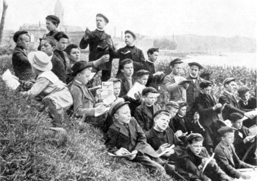Tekenles op de Scheldedijk. Aan de overzijde van de stroom ligt Temse, met de sierlijke toren van de Onze-Lieve-Vrouwekerk en met de schoorstenen van de fabrieken waar veel inwoners in zware omstandigheden hun dagelijks brood moes-ten verdienen. Edward is de derde van links, boven het jongetje met de strohoed. 